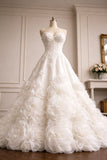 A strapless white wedding gown with a sweetheart neckline, fitted lace bodice, and a voluminous skirt made of layered fabric roses, displayed on a mannequin in an elegant indoor room.