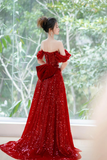 Woman in a red off-shoulder evening gown standing in front of a window with greenery.