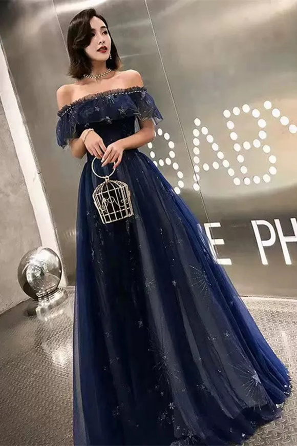 Woman in a blue off-shoulder dress holding a birdcage handbag in an indoor setting.