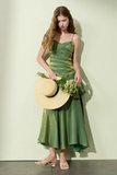 Woman in a green dress holding a straw hat and flowers against a plain background