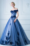 Woman wearing a blue evening gown with floral embellishments against a white wall.