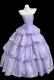 A lavender purple strapless ball gown displayed on a white mannequin torso against a solid black background. The dress features a structured bodice, a large ruffled tiered skirt, and a matching fabric flower detail at the waist.