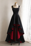 A sleeveless black formal gown displayed on a dress form, featuring a high-low layered skirt with a deep red lining, set against a neutral indoor background with a white curtain.