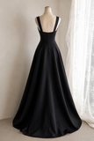 A floor-length black satin gown displayed on a dress form mannequin, featuring a low scoop neckline, wide shoulder straps, and a full A-line skirt, set against a neutral indoor background with a white lace curtain.