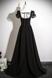 A long black formal gown displayed on a mannequin against a white wall. The dress has sheer puff sleeves with black lace inserts, a square neckline with a front tie, and a full floor-length skirt.