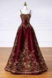 A floor-length burgundy satin ball gown with intricate gold floral embroidery, displayed on a white mannequin against a white paneled wall and wooden floor.