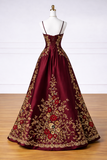 A floor-length burgundy satin gown with gold floral embroidery and red rose embellishments, displayed on a white mannequin against a white paneled wall and wood floor.