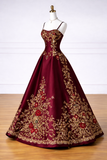 A floor-length burgundy ball gown with gold floral embroidery and red rose motifs, displayed on a white mannequin against a white paneled wall and light wood floor.