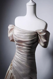 A champagne-colored off-the-shoulder evening gown with a shimmering satin-like finish, displayed on a white mannequin torso against a dark gray gradient background.