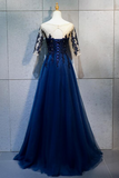 A floor-length navy blue tulle evening gown displayed on a mannequin against a gray tiled wall. The dress has sheer off-the-shoulder sleeves with dark floral lace applique, a sweetheart neckline, and a corset-style bodice with lace and bead detailing.