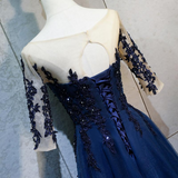 A navy blue formal gown displayed on a cream mannequin, featuring sheer off-the-shoulder sleeves with dark navy floral lace applique, a lace-up corset-style bodice, and a fitted skirt.
