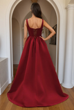 A woman wearing a floor-length burgundy A-line gown, photographed from the back in an elegant indoor hallway with white arches and columns.