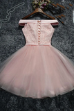 A light pink off-the-shoulder tulle dress with lace-up back detailing, displayed on a wooden hanger over a dark gray textured rug with decorative props.