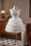 A white lace corset-style dress displayed on a mannequin in an indoor room. The dress has thin shoulder ties, a fitted lace-up bodice, and a layered ruffled tulle skirt.