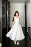 A woman wearing a sleeveless white midi dress with a square neckline and a voluminous tulle skirt, standing in a hallway with dark floors and mirrored walls.