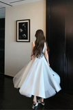 A woman viewed from behind wearing a sleeveless white A-line midi dress with a sheer tulle hem, standing in a room with dark wood floors and a beige wall.