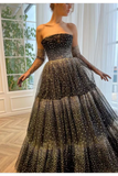 A strapless black tulle ball gown with sheer long sleeves, covered in shimmering gold polka dots, worn by a model in a bright room with large windows.