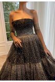 A strapless black tulle ball gown with sheer long sleeves, covered in gold polka dots, worn by a model in an indoor setting.