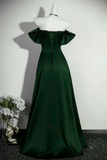 A floor-length dark green satin gown displayed on a white mannequin, featuring an off-the-shoulder neckline, ruffled sleeves, a lace-up back, and a full skirt. The mannequin is set against a plain light wall with a decorative vase of white flowers on the floor to the left.