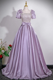 A lavender purple formal ball gown displayed on a mannequin. The dress features a square neckline with a heavily embellished, beaded bodice, short puff sleeves with bow details at the shoulders, a matching bow at the waist, and a full, flowing skirt.