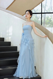 A woman wearing a light blue strapless gown with a ruffled tiered skirt, standing on a black staircase next to a curved cream-colored wall.