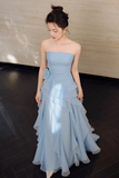 A woman wearing a strapless light blue gown with a tiered ruffled skirt, standing in an indoor setting with a dark wood floor and a light beige wall.