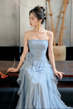 A woman wearing a light blue strapless gown with a ruffled tiered skirt, seated indoors near a wooden table and potted plants.