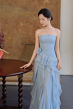 A woman wearing a strapless light blue gown with a ruffled tiered skirt, standing beside a dark wooden table in an indoor setting with a textured wall background.