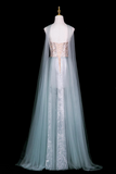 A long flowing gown displayed on a mannequin against a black background. The dress has a pale icy blue sheer tulle overlay, a white underlayer with silver floral embroidery, and a beige corset-style bodice with gold lace-up detailing.