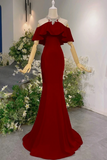 A deep red off-the-shoulder mermaid gown displayed on a mannequin with wooden arms, set against a white wall decorated with green foliage and white flowers.