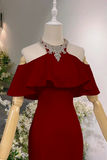 A deep red off-the-shoulder evening gown displayed on a cream mannequin torso, styled with a sparkling silver crystal choker necklace. The background features a soft gray wall and decorative white floral arrangements.