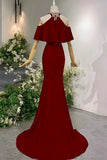 A deep red off-the-shoulder mermaid gown displayed on a mannequin in an indoor showroom setting with decorative white flowers and greenery.