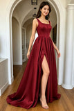 A woman models a floor-length burgundy satin gown with a square neckline, fitted bodice, and high thigh slit, standing in a white arched hallway with wood floors.